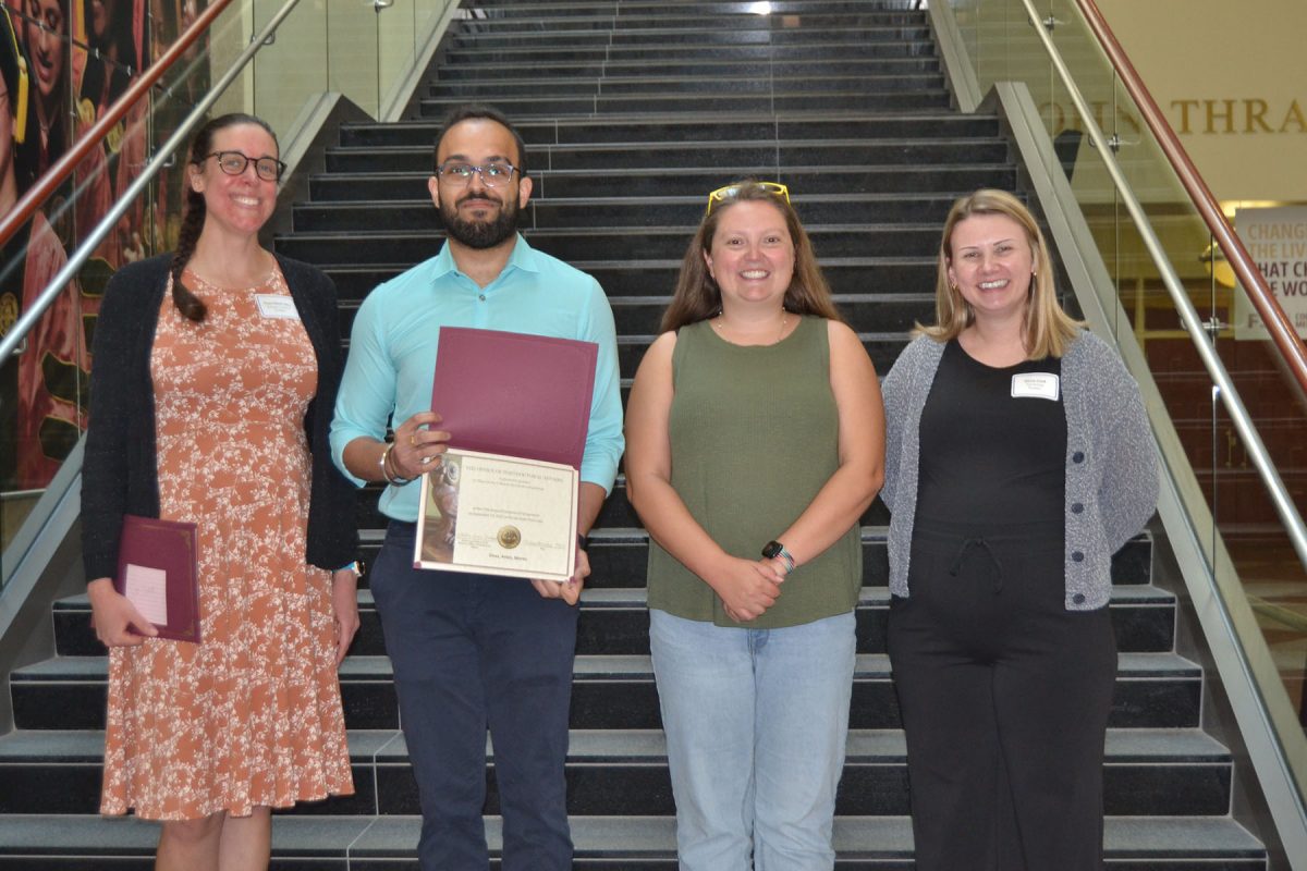 Four people pose for a photo in front of stairs, one of them holding a piece of paper looking to be a certificate of some kind