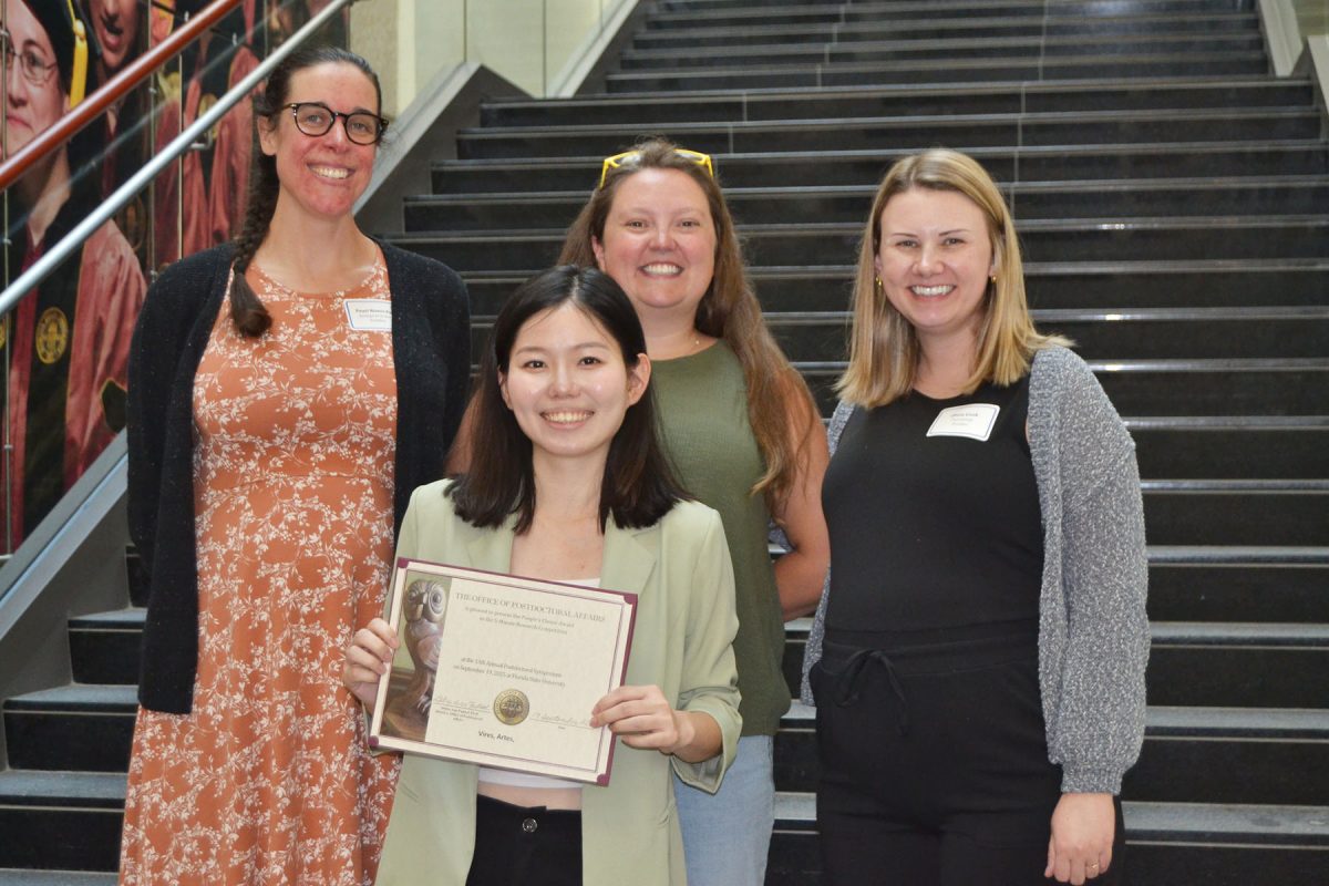 Four people smile and pose for a photo in front of stairs. One of them is standing in front of the other three, holding a piece of paper looking to be a certificate of some kind