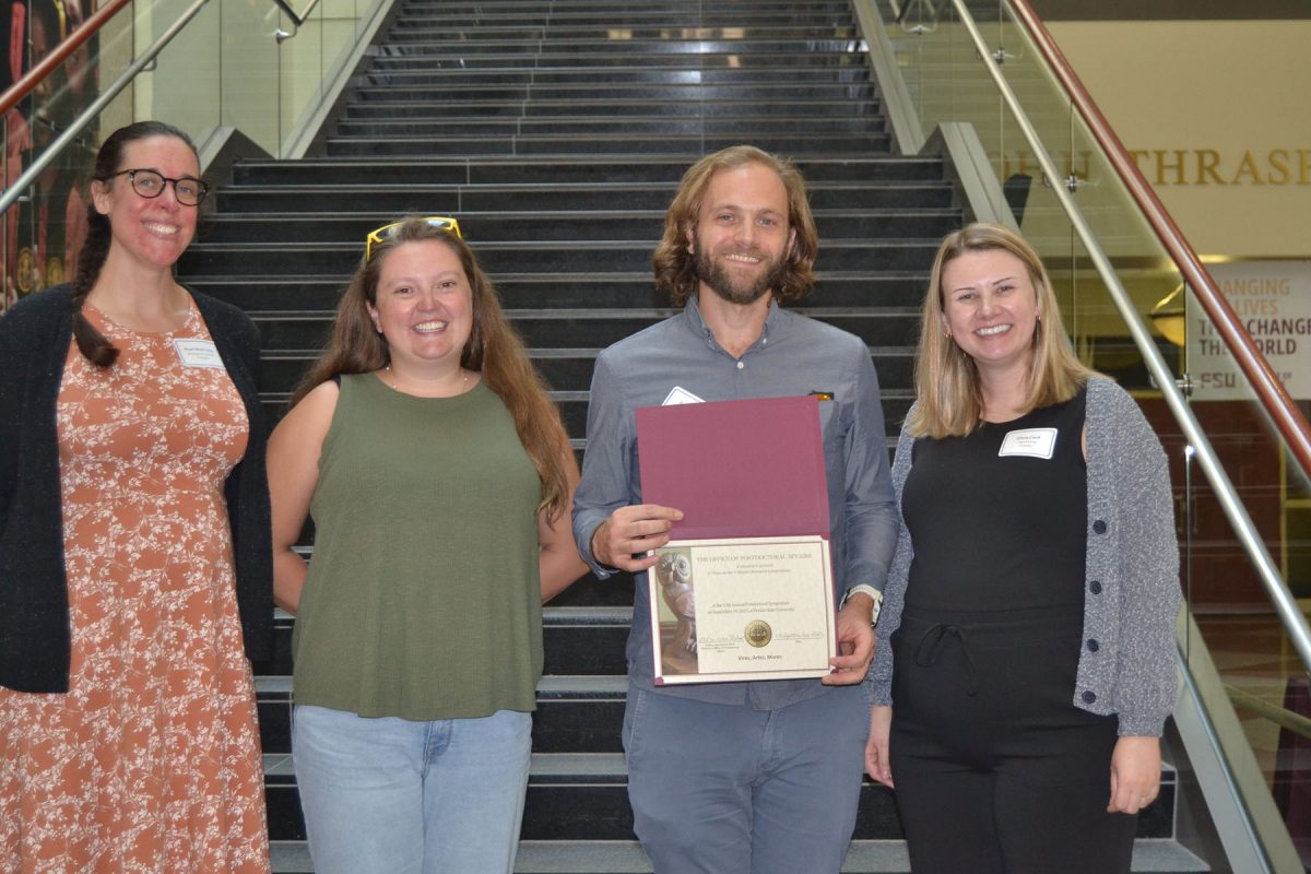 Four people smile and pose for a photo in front of stairs, one of them holding a certificate.