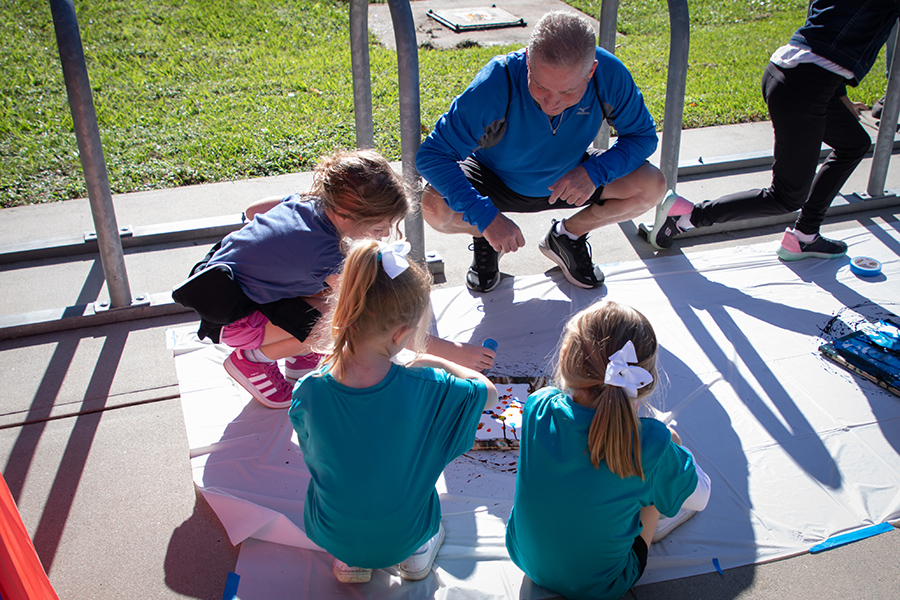 One man crouches down next to three children painting